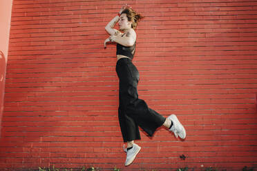 Woman jumping against red brick wall - MEUF01711