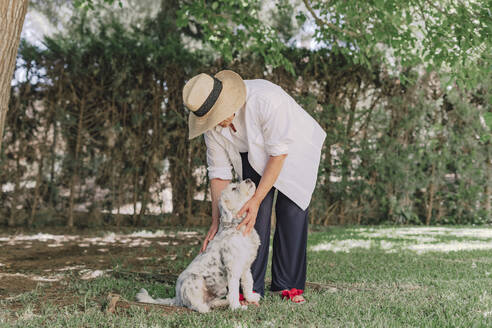 Senior woman wearing hat with dog standing on grassy land in yard - ERRF04118