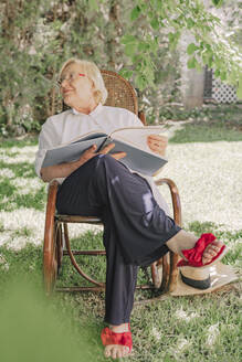 Senior woman holding book looking away while relaxing on chair in yard - ERRF04119