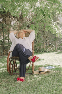 Thoughtful senior woman with hands behind head relaxing on chair in yard - ERRF04121