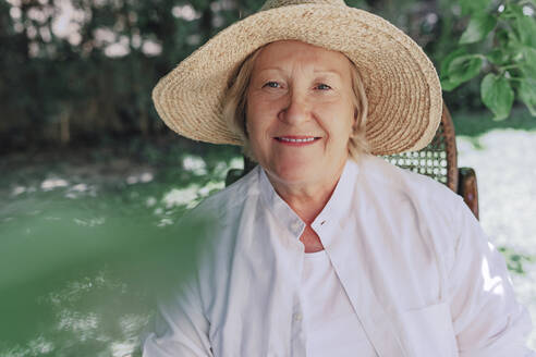 Close-up of smiling senior woman wearing hat sitting on chair in yard - ERRF04123