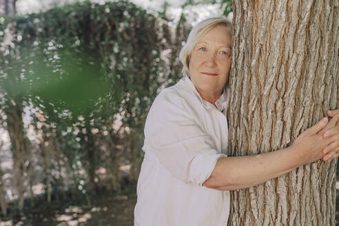 Senior woman embracing tree trunk while standing in yard - ERRF04124