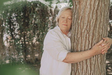Ältere Frau mit geschlossenen Augen umarmt einen Baumstamm, während sie im Hof steht - ERRF04125