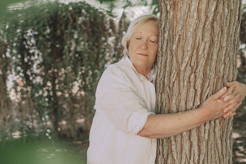 Senior woman with eyes closed embracing tree trunk while standing in yard - ERRF04125