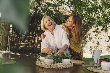 Tochter schaut auf fröhliche Mutter, die sich um Pflanzen auf einem Tisch im Garten kümmert - ERRF04139