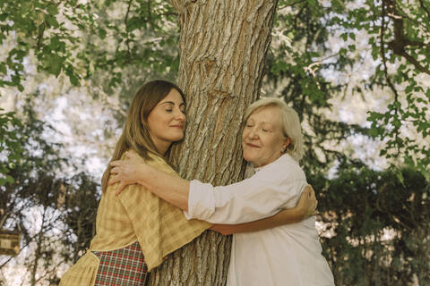 Mother and daughter with eyes closed embracing tree trunk in yard stock photo