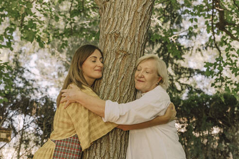 Mother and daughter with eyes closed embracing tree trunk in yard - ERRF04150