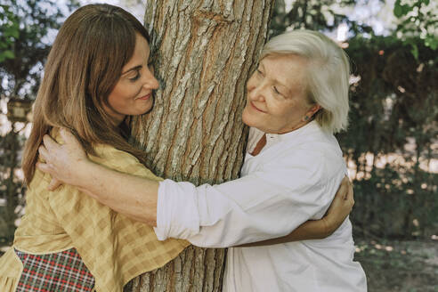 Mother and daughter looking at each other while embracing tree trunk in yard - ERRF04151