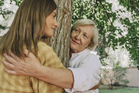 Mother looking at daughter while embracing tree trunk with her in yard stock photo