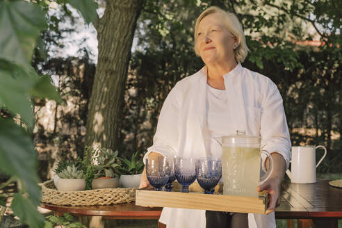 Thoughtful senior woman carrying tray with drinks while standing in yard - ERRF04158