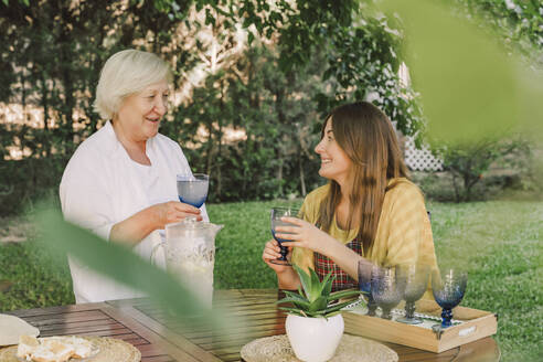 Smiling mother and daughter enjoying drinks while talking in yard - ERRF04159