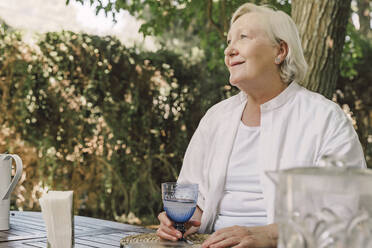 Nachdenkliche ältere Frau hält ein Getränk in der Hand, während sie an einem Tisch vor Bäumen im Hof sitzt - ERRF04162