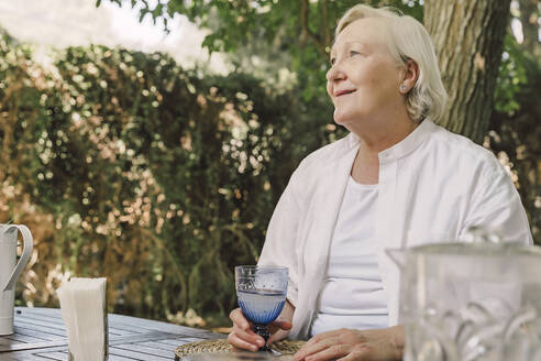 Thoughtful senior woman holding drink while sitting at table against trees in yard - ERRF04162