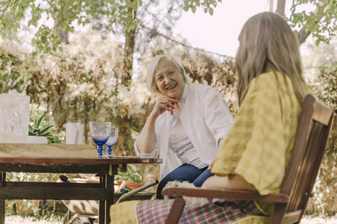 Happy senior woman talking with daughter while sitting at table in yard - ERRF04164