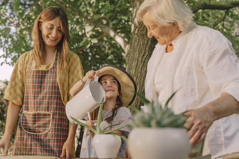 Smiling mother and grandmother looking at girl watering potted plant in yard - ERRF04172