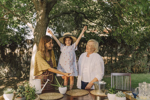 Cheerful girl with arms raised standing with mother and grandmother against tree in yard stock photo