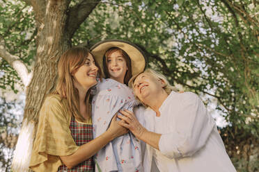 Happy family embracing girl wearing hat against tree in yard - ERRF04174
