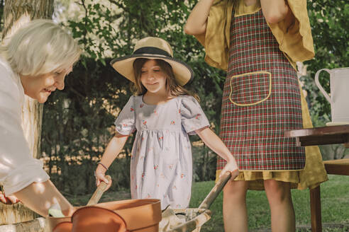 Girl holding wheelbarrow while standing with mother and grandmother in yard - ERRF04175