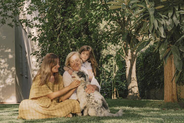 Multi-generation family playing with dog on grassy land against plants in yard - ERRF04178