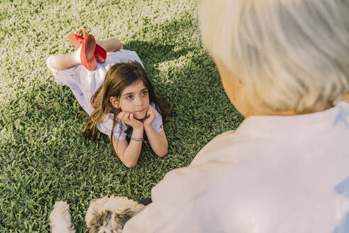 Cute girl with hands on chin looking at grandmother while lying over grassy land in yard - ERRF04179