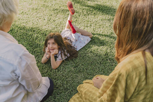Cute girl with hands on chin talking with family while lying over grassy land in yard - ERRF04180