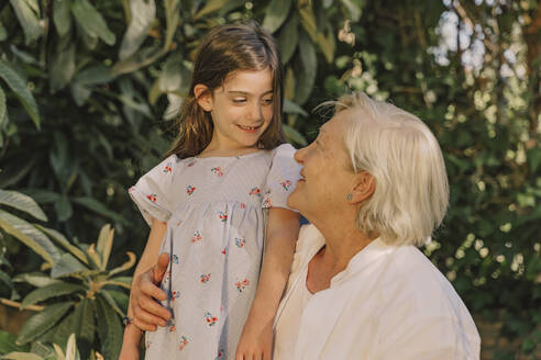 Lächelnde ältere Frau schaut auf süße Enkelin gegen Pflanzen im Hof - ERRF04182