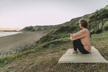 Nachdenkliche junge Frau sitzt auf einer Gymnastikmatte am Strand gegen den klaren Himmel bei Sonnenuntergang - MTBF00566