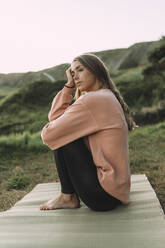 Thoughtful woman sitting on exercise mat at beach against clear sky during sunset - MTBF00567