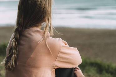 Nahaufnahme einer jungen Frau mit geflochtenem Haar, die bei Sonnenuntergang am Strand sitzt - MTBF00568