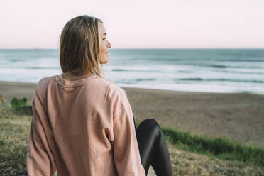 Junge Frau schaut bei Sonnenuntergang am Strand sitzend aufs Meer - MTBF00569