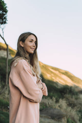 Thoughtful young woman with arms crossed standing at beach against clear sky during sunset stock photo