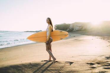Junge Frau im Neoprenanzug mit Surfbrett am Strand bei Sonnenuntergang - MTBF00572