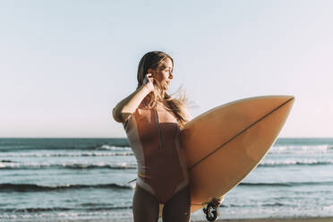 Junge Frau mit Surfbrett, die bei Sonnenuntergang auf dem Meer steht und wegschaut - MTBF00573