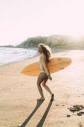 Fröhliche junge Frau mit Surfbrett am Strand gegen den klaren Himmel bei Sonnenuntergang - MTBF00574