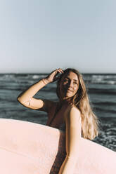 Junge Frau mit Surfbrett, die wegschaut, während sie auf dem Meer steht - MTBF00580