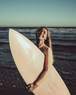 Cheerful woman with eyes closed carrying surfboard against sea during sunset - MTBF00581