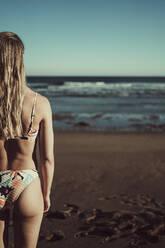 Junge Frau im Bikini, die bei Sonnenuntergang am Strand steht und aufs Meer schaut - MTBF00585