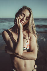 Nahaufnahme einer jungen Frau mit geschlossenen Augen im Bikini, die bei Sonnenuntergang am Meer steht - MTBF00587