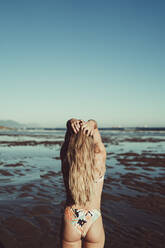 Junge Frau mit Händen in den Haaren steht am Strand gegen klaren Himmel - MTBF00591