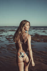 Young woman in bikini looking away while walking at beach against clear sky during sunset - MTBF00592