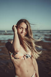 Smiling young woman in bikini standing against sea and clear sky during sunset - MTBF00593