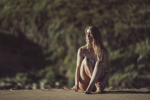 Nachdenkliche junge Frau im Bikini am Strand sitzend bei Sonnenuntergang - MTBF00595