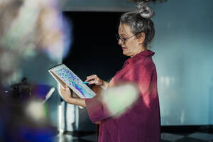 Senior woman wearing eyeglasses drawing in book while standing at home - ERRF04205