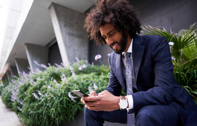 Joyful ethnic man in suit sitting outside and messaging - ADSF08256