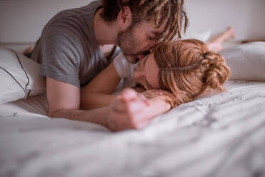Zärtlicher Mann mit Dreadlocks umarmt und küsst Frau mit roten Haaren, während sie zusammen auf dem Bauch auf dem Bett liegen und sich am Wochenende entspannen - ADSF08268