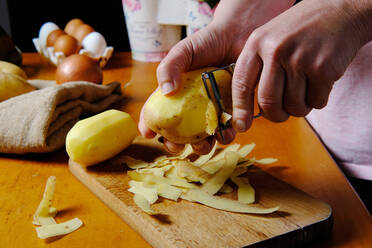 Crop anonymous cook peeling potato above wooden cutting board in modern kitchen - ADSF08318