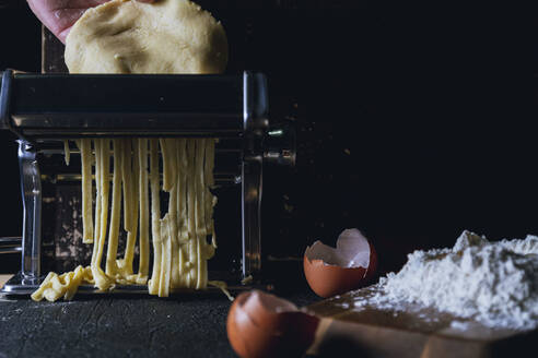 Crop Person bereitet hausgemachte Fettuccine mit Nudelmaschine auf dem Tisch mit Mehl und Eierschale gegen schwarzen Hintergrund platziert - ADSF08329