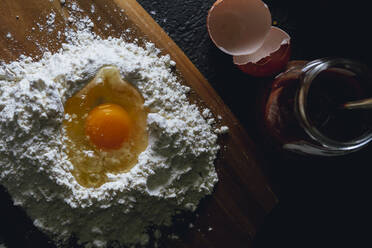 Top view of broken egg in flour on textured black surface with round knife for cutting dough and glass pot - ADSF08330