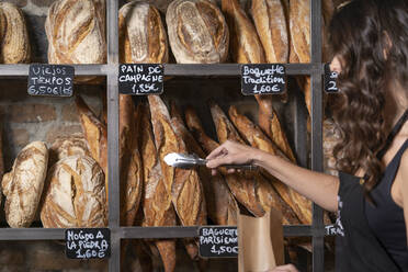 Unbekannte Frau in schwarzer Schürze legt französisches Baguette in Papiertüte mit Metallbändern in Bäckerei - ADSF08373