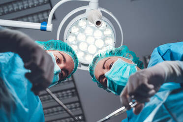 From below female surgeons in medical uniform using professional tools while standing under bright light in operating theater - ADSF08417